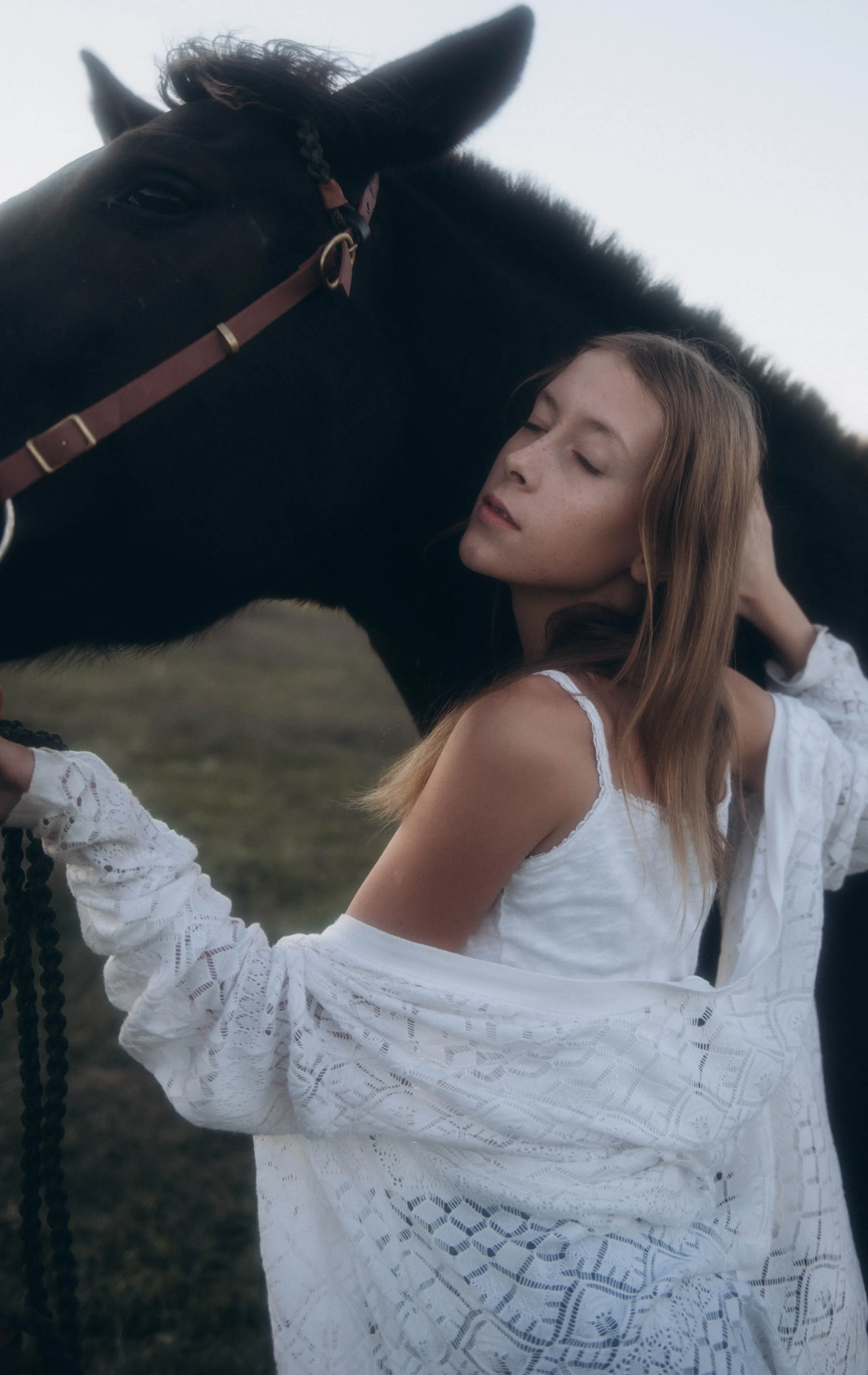 A serene scene of a young woman in white, embracing a black horse in a rural setting.