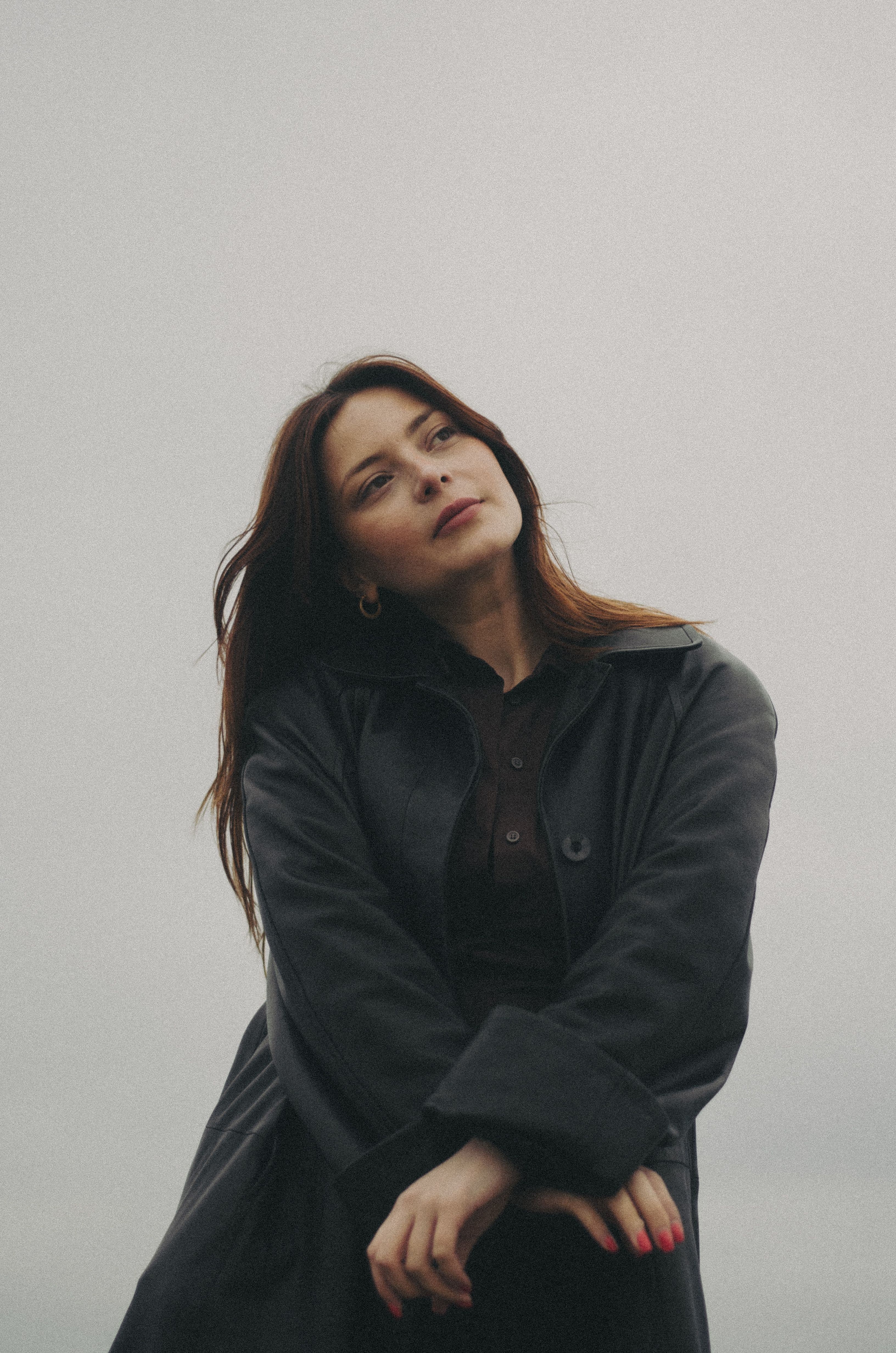 A thoughtful woman poses elegantly in a black coat against a neutral background.