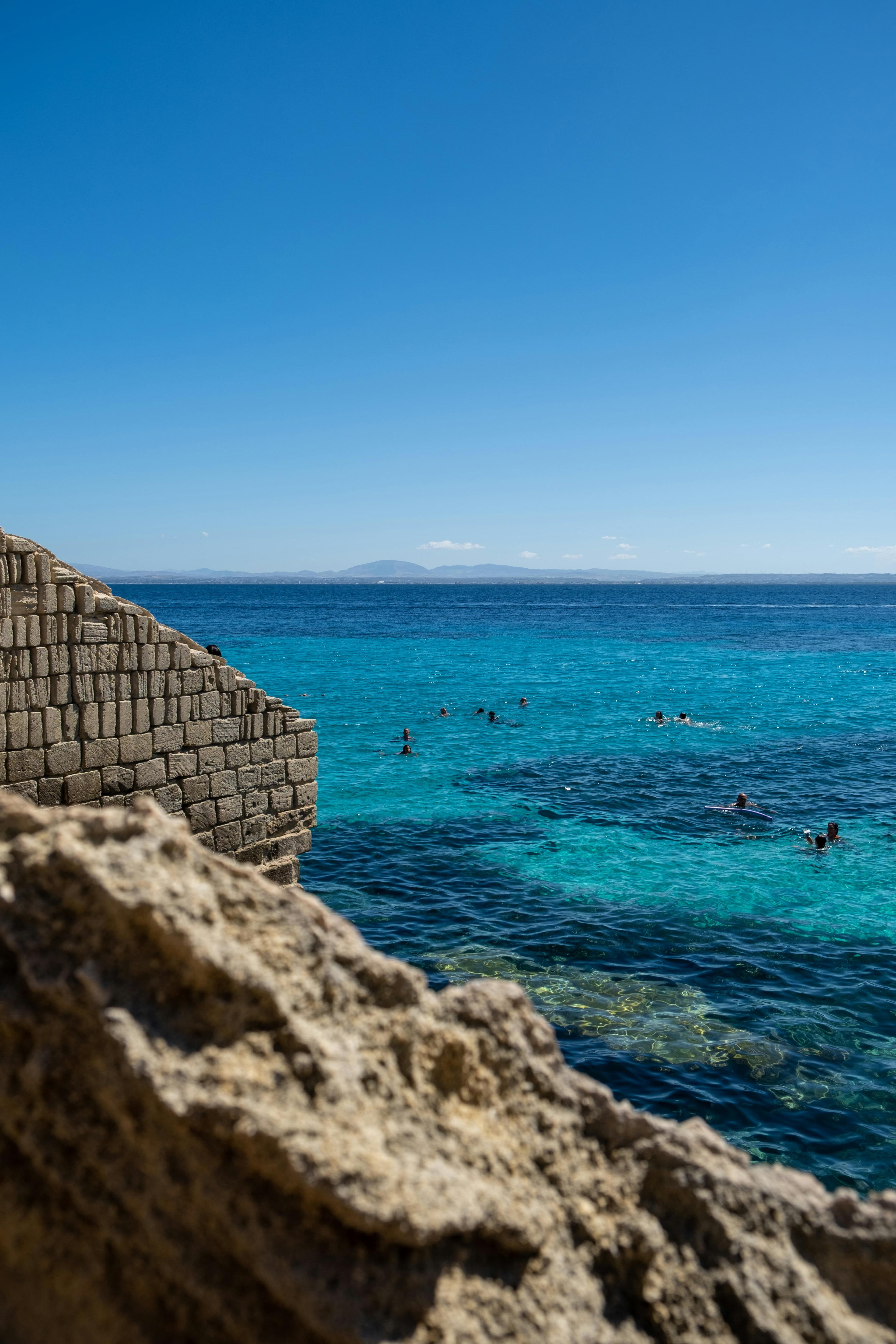 Clear turquoise waters with tourists swimming near a stone structure on a sunny day.