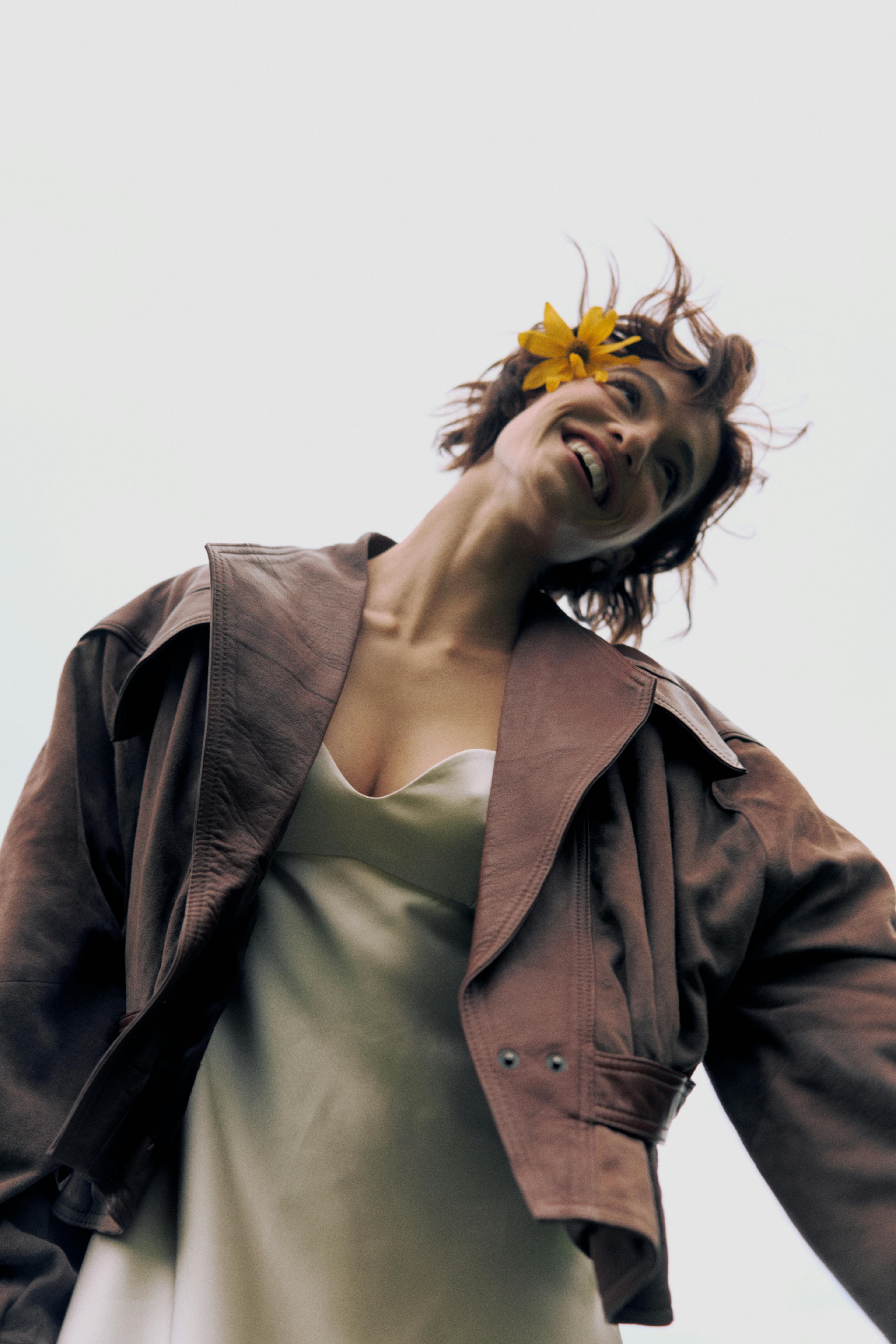 Woman outdoors with a bright smile, wearing a leather jacket and flower in hair, showcasing carefree fashion.