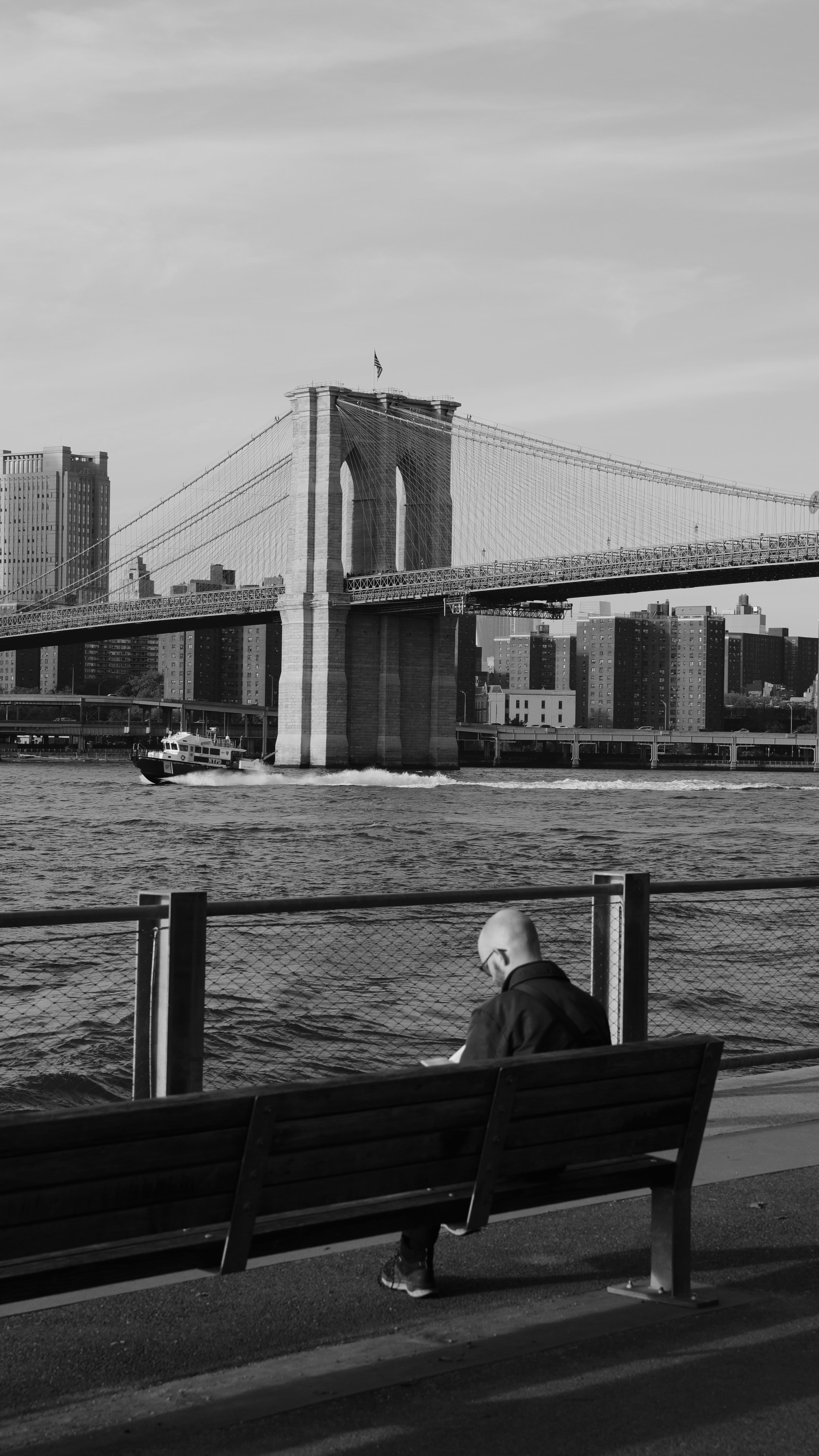 Bald man sits on a bench gazing at the Brooklyn Bridge in this black and white cityscape.