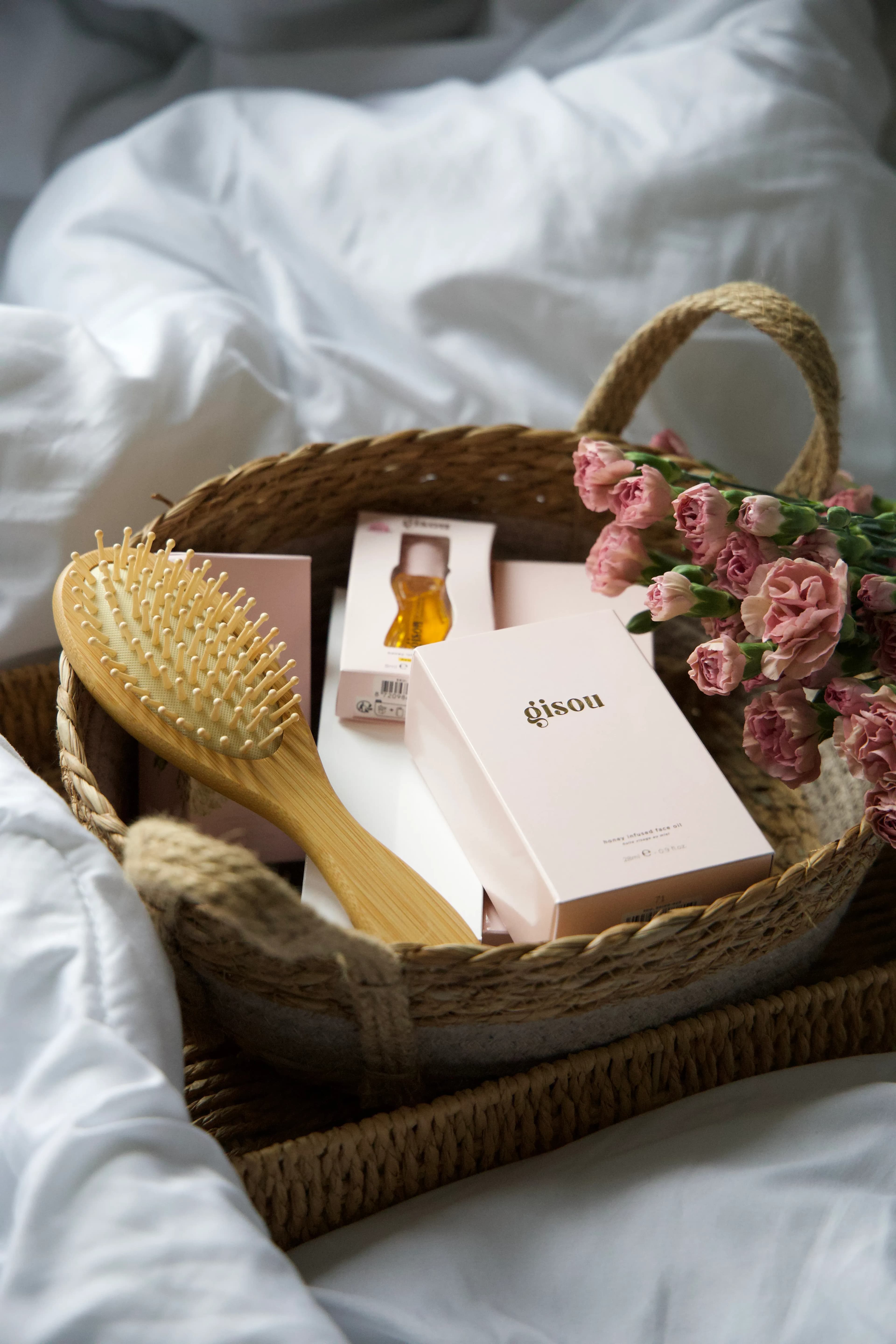 Woven basket with haircare products, hairbrush, and pink flowers on white bedding.