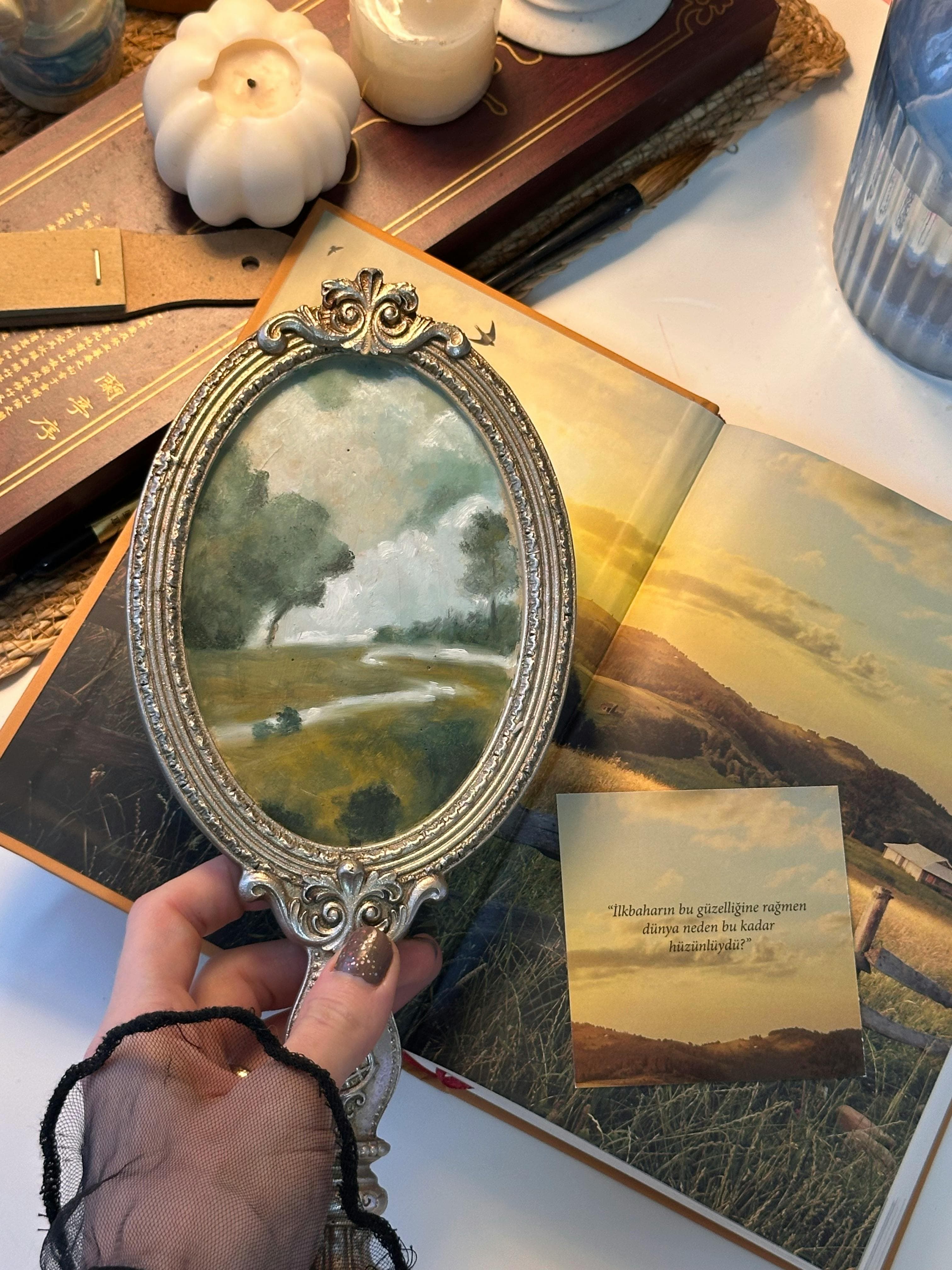 A vintage oval mirror held over an open book reveals a pastoral painting. Elegant decor setup.