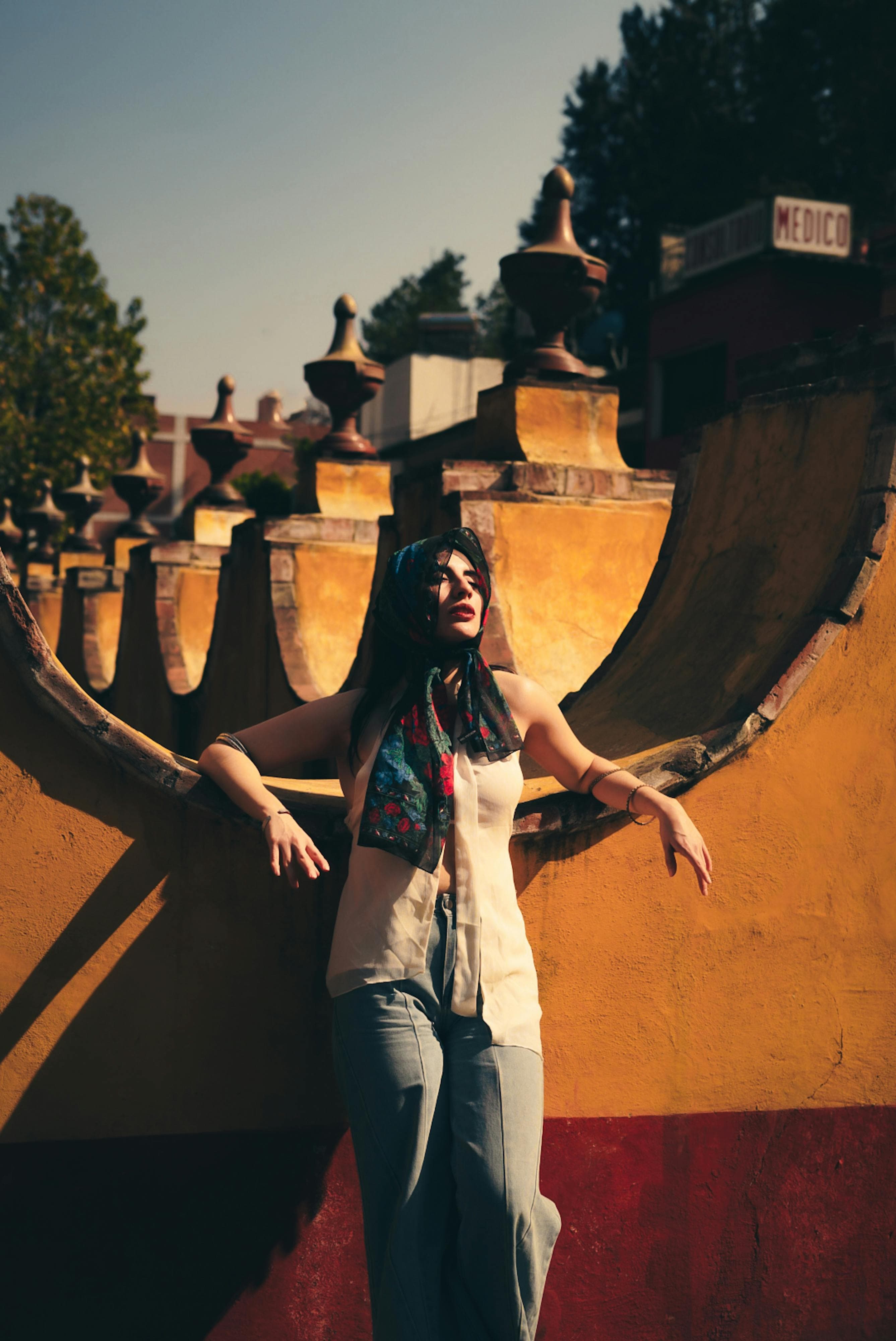 A woman in casual attire leans against a vibrant yellow wall under the warm sunlight.